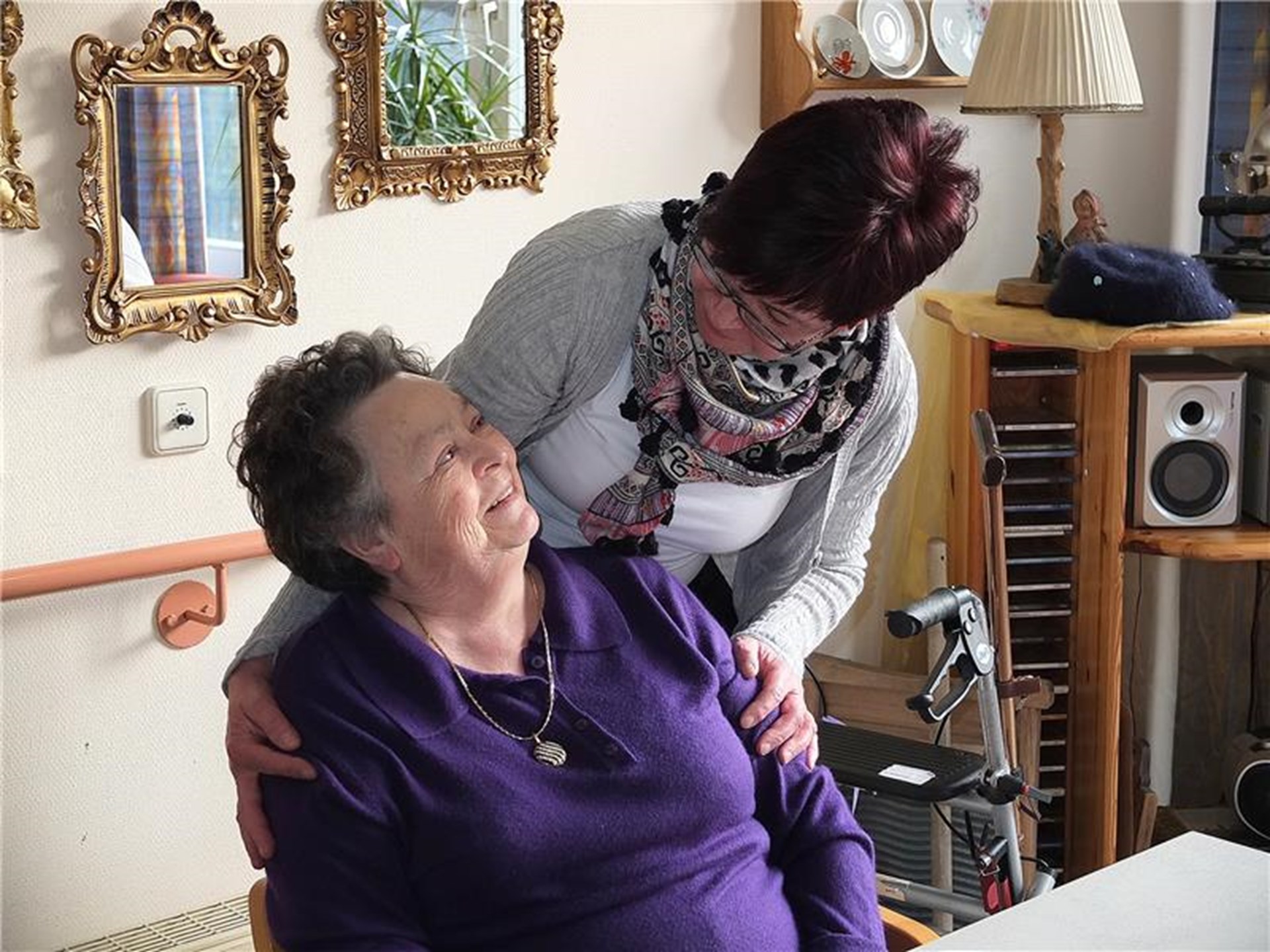 Eine Bewohnerin sitzt auf einem Stuhl in ihrem Zimmer. Hinter ihr steht eine jüngere Frau und legt ihr die Hände auf die Schultern. Die zwei Frauen lächeln sich an. Im Hintergrund steht ein Regal, ein Rollator und Bilder hängen an den Wänden.