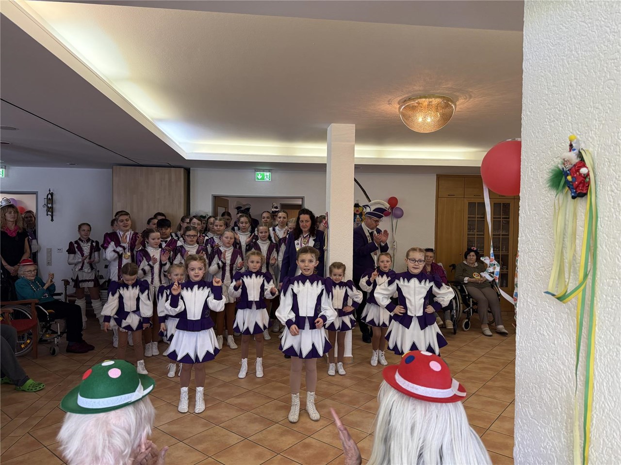 In einem Kreis sitzen Bewohner:innen mit Karnevalshüten. In der Mitte steht eine große Karnevalsgruppe oder Garde in Kostümen. Eine Gruppe junger Mädchen tanzt.