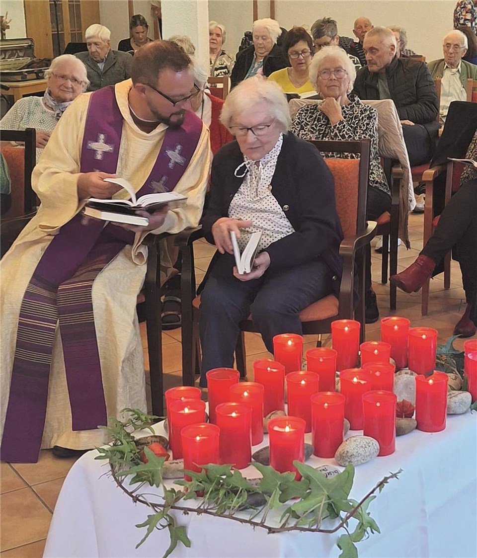 Die Bewohner:innen haben sich zu einem Gottesdienst in der Kapelle versammelt. Der Pfarrer schaut mit einer Bewohnerin in ein Gebetsbuch. Im Vordergrund stehen viele rote Kerzen auf einem Tisch.