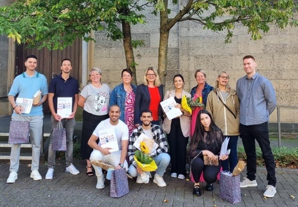 Eine Gruppe von Menschen, darunter mehrere Personen, die erfolgreich ihre Ausbildung beendet haben, stehen vor einer Kirche. Sie haben ihre Zeugnisse, Blumen und Geschenktüten in der Hand.