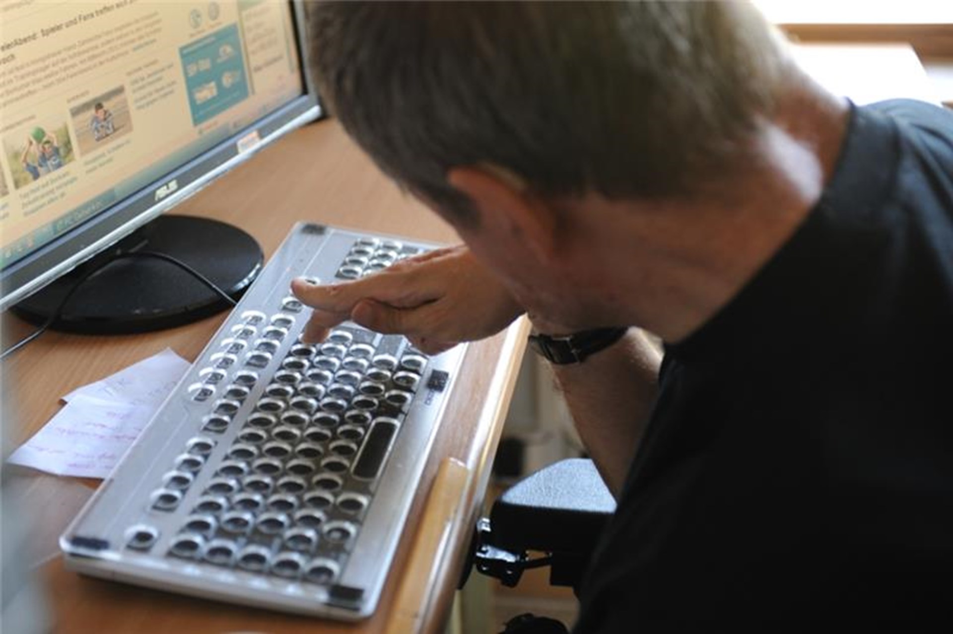 Blick über die Schulter eines Mannes mit Beeinträchtigung, der am Computer sitzt und die Tastatur benutzt.