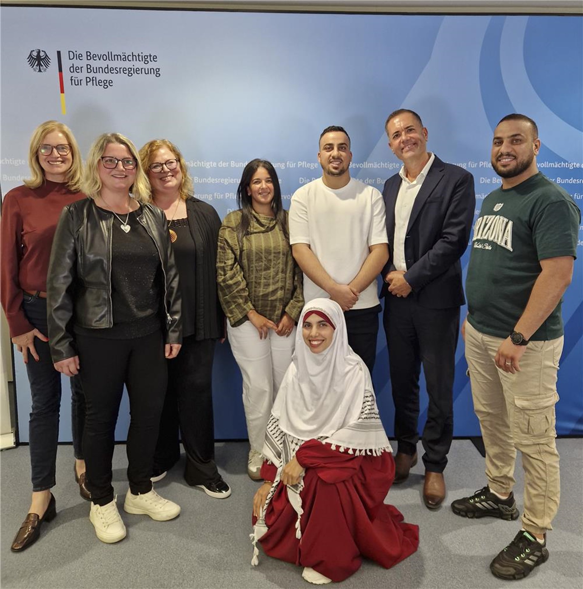 Gruppenfoto von acht lächelnden Personen vor einer blauen Pressewand mit dem Schriftzug „Die Bevollmächtigte der Bundesregierung für Pflege“ und dem Bundesadler. Sieben Personen stehen nebeneinander, während eine Frau mit weißem Kopftuch und rotem Kleid vorne kniet.