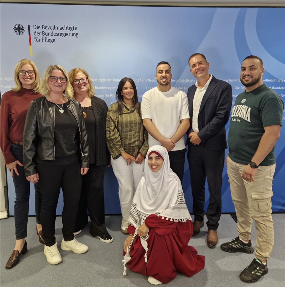 Gruppenfoto von acht lächelnden Personen vor einer blauen Pressewand mit dem Schriftzug „Die Bevollmächtigte der Bundesregierung für Pflege“ und dem Bundesadler. Sieben Personen stehen nebeneinander, während eine Frau mit weißem Kopftuch und rotem Kleid vorne kniet.
