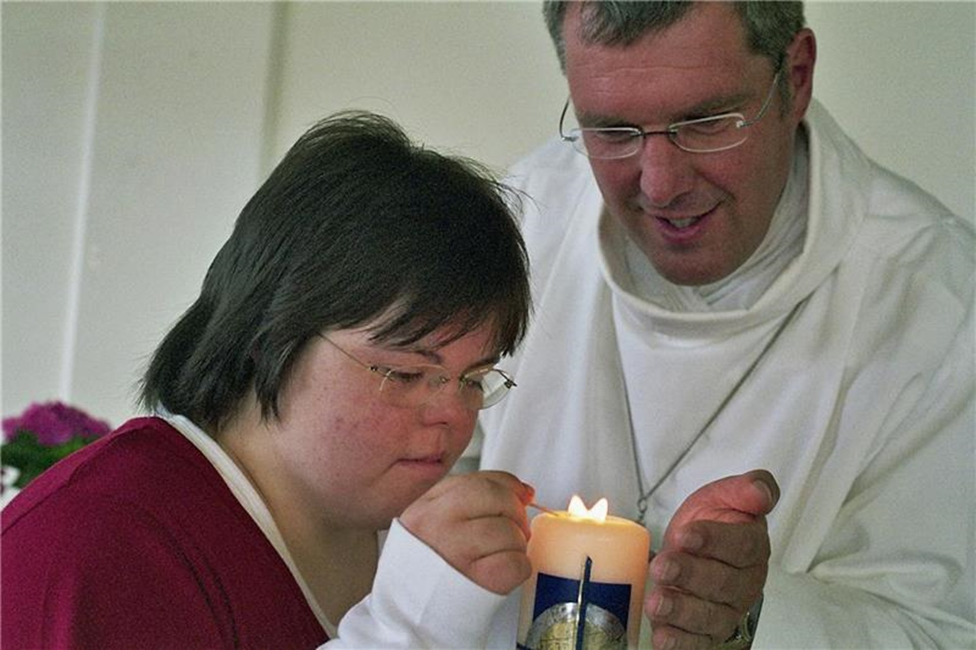 Ein Priester und eine Frau mit Downsyndrom zünden gemeinsam eine Kerze an.