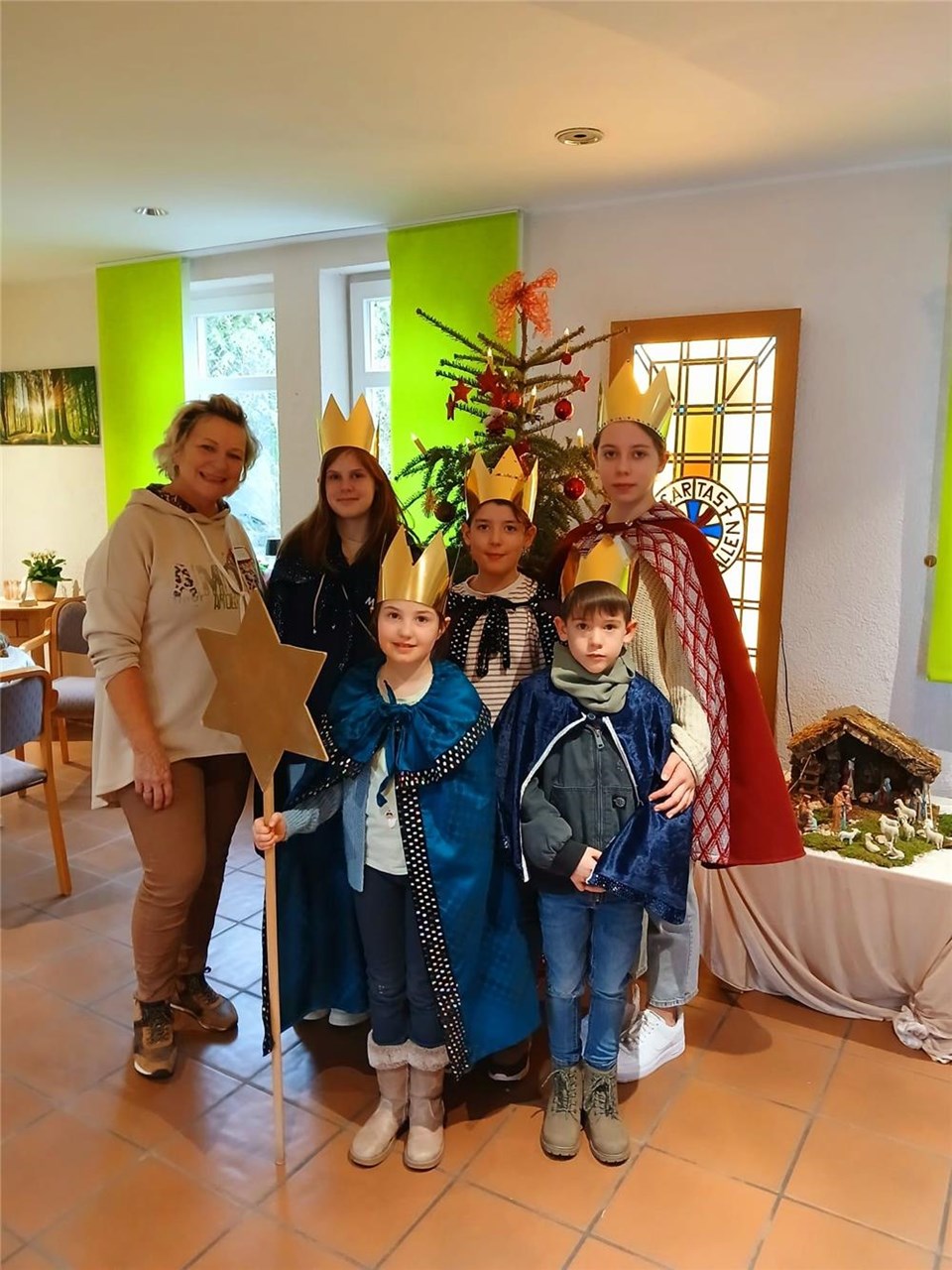 5 Mädchen und Jungen sind als Sternsinger mit Kronen auf den Köpfen verkleidet. Sie stehen mit einer Mitarbeiterin vor einem Tannenbaum.
