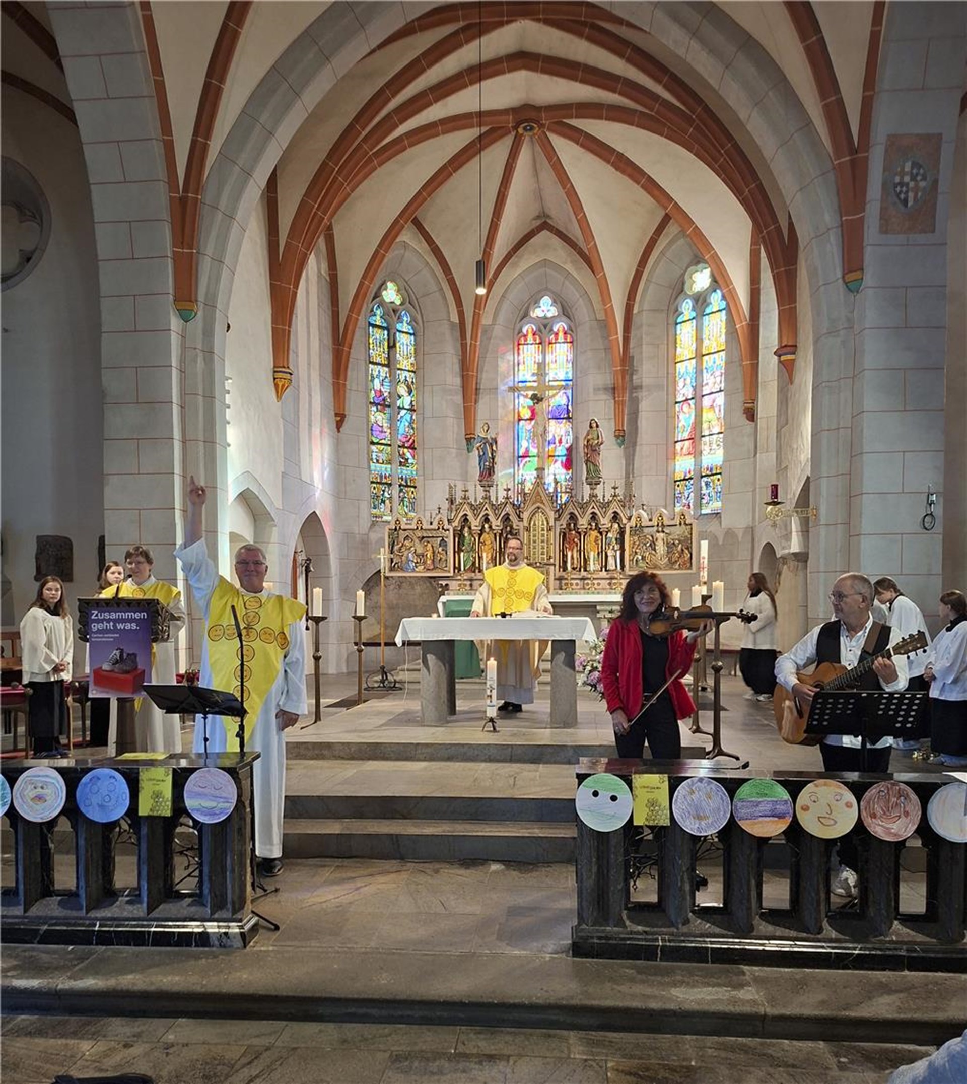 Blick in eine Kirche mit farbigen Buntglasfenstern und reich verziertem Altar. Vor dem Altar stehen Geistliche und Musiker mit Geige und Gitarre während Kinderzeichnungen die Absperrung im Vordergrund schmücken.