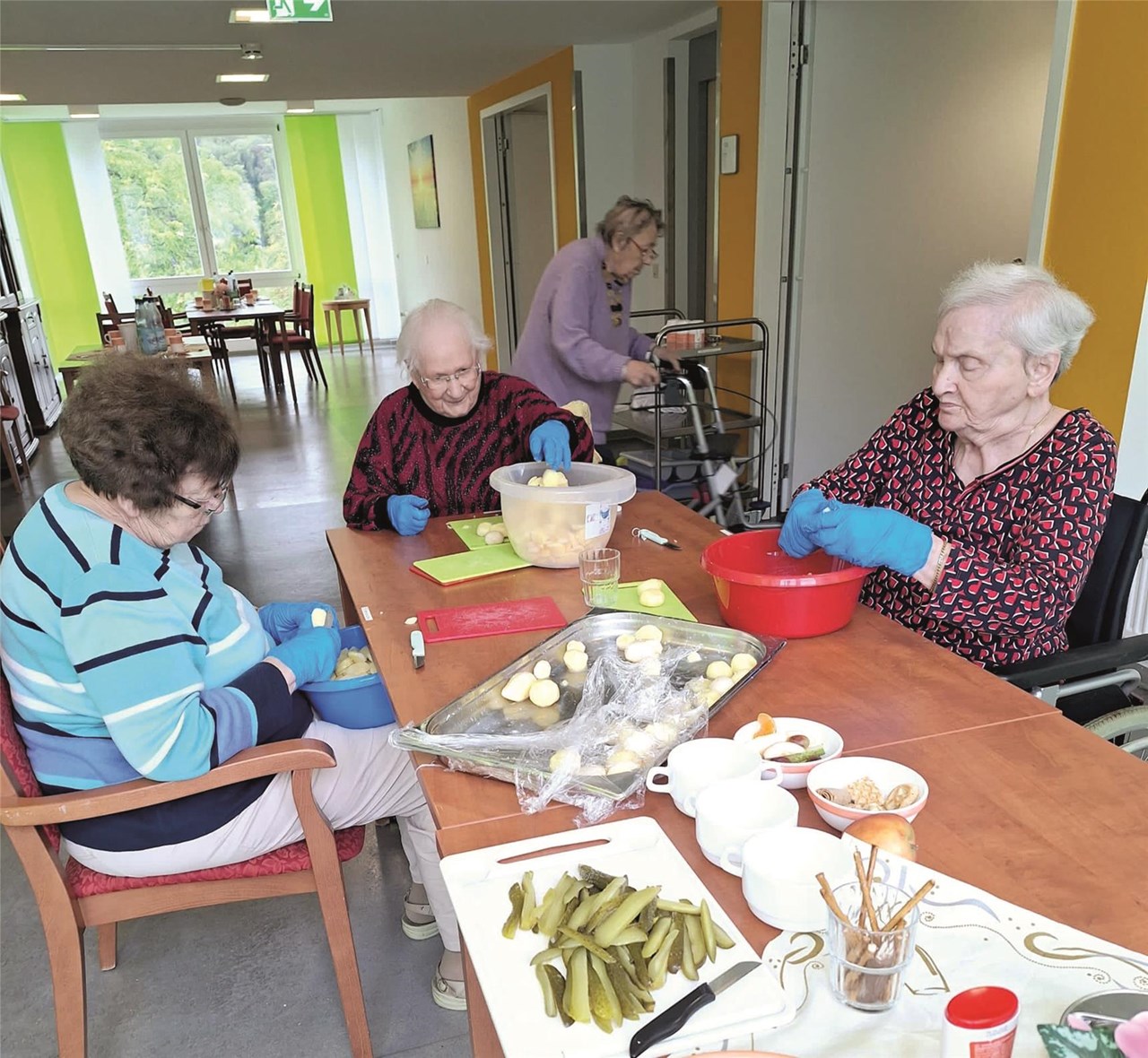 Drei Bewohnerinnen sitzen an einem Tisch und schneiden Kartoffeln in Scheiben. Auf dem Tisch steht eine Platte mit geschnittenen Essiggurken.