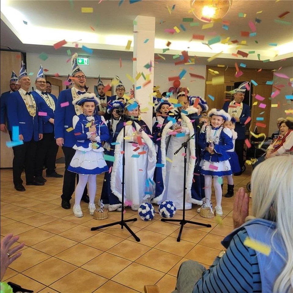 Eine Karnevalsgruppe mit blau-weißen Kostümen singt und klatscht. Vier Mädchen benutzen Konfettikanonen. Konfetti fliegt durch die Luft. Es wird gelacht und geklatscht.