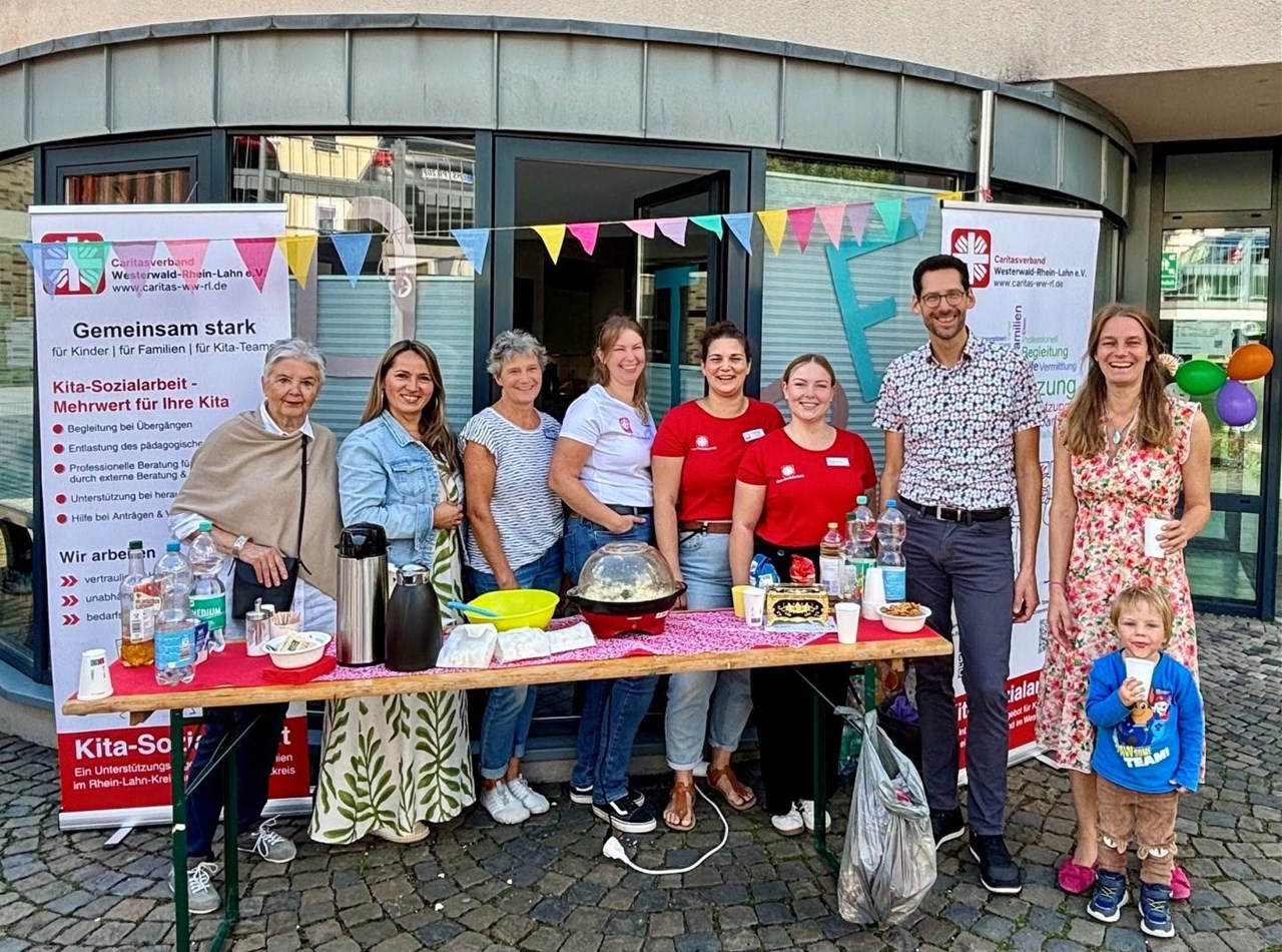 Acht Erwachsene und ein kleines Kind stehen lächelnd an einem Getränkestand der Caritas-Kita-Sozialarbeit. Auf dem Tisch stehen Getränke und Snacks. Im Hintergrund sind Informationsbanner der Caritas und eine bunte Wimpelkette zu sehen.