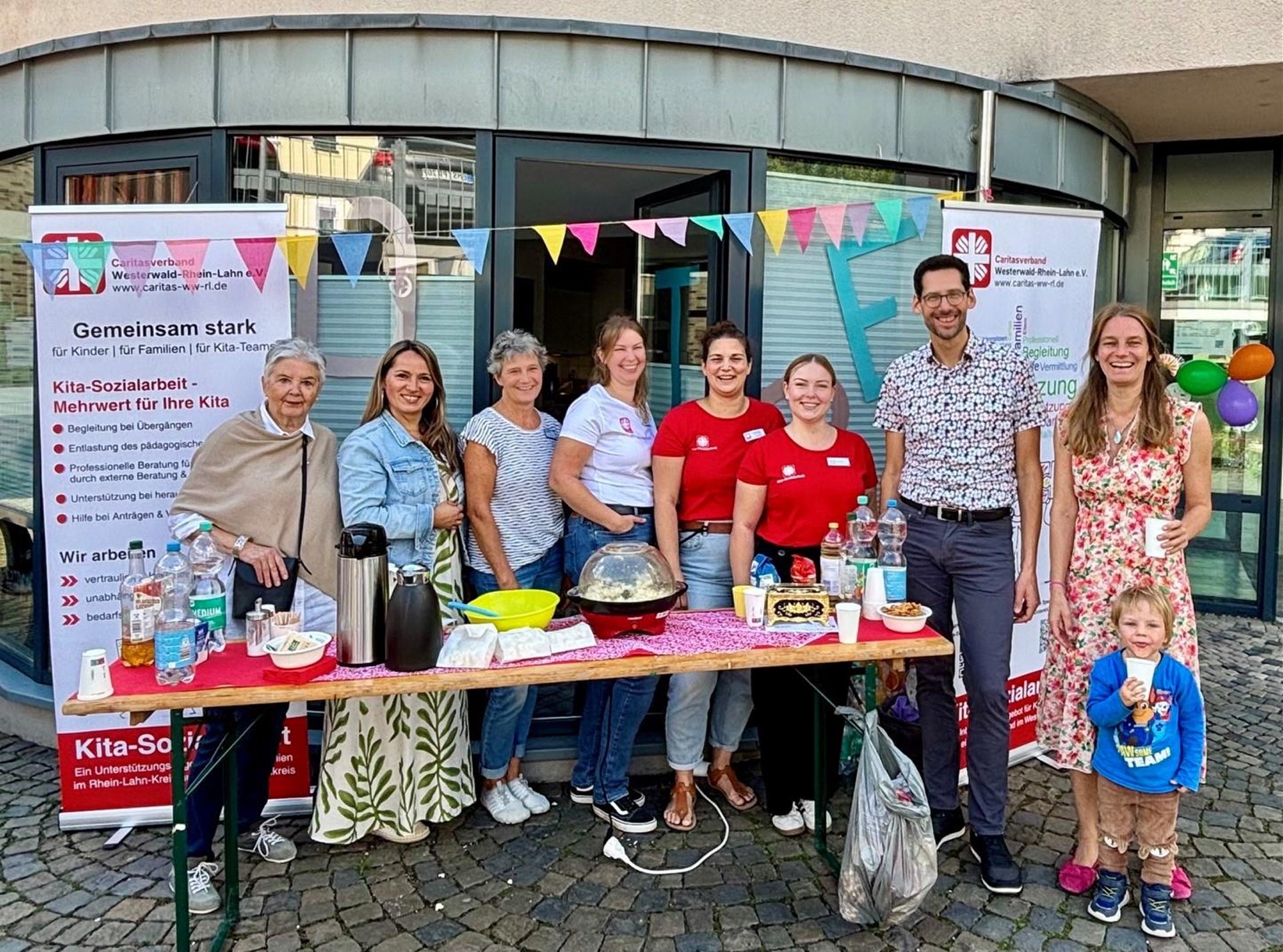Acht Erwachsene und ein kleines Kind stehen lächelnd an einem Getränkestand der Caritas-Kita-Sozialarbeit. Auf dem Tisch stehen Getränke und Snacks. Im Hintergrund sind Informationsbanner der Caritas und eine bunte Wimpelkette zu sehen.