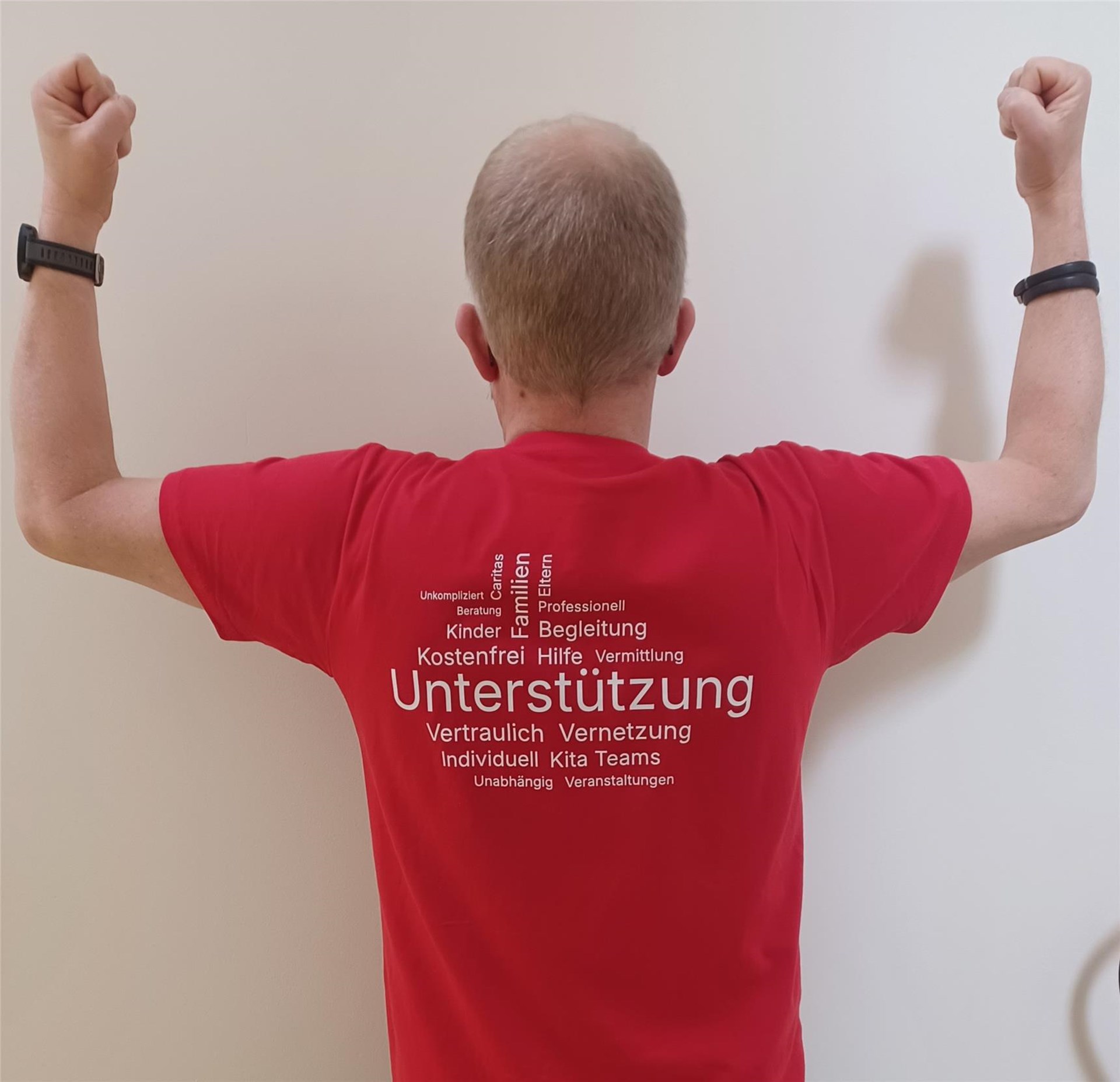 Person in rotem T Shirt steht mit dem Rücken zur Kamera und hebt beide Arme angewinkelt nach oben. Auf dem Shirt stehen die Wörter „Unterstützung“, „Vertraulich“, „Vernetzung“, „Individuell“, „Professionell“, „Begleitung“, „Familien“, „Kinder“, „Hilfe“ und weitere Begriffe in weißer Schrift.