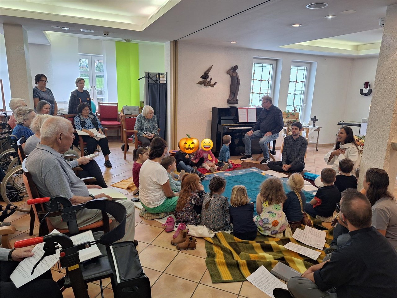 Eine Gruppe von Kindern sitzt auf Decken in einem Kreis auf dem Boden. Um sie herum sitzen auf Stühlen die Bewohner:innen. Sie singen. 