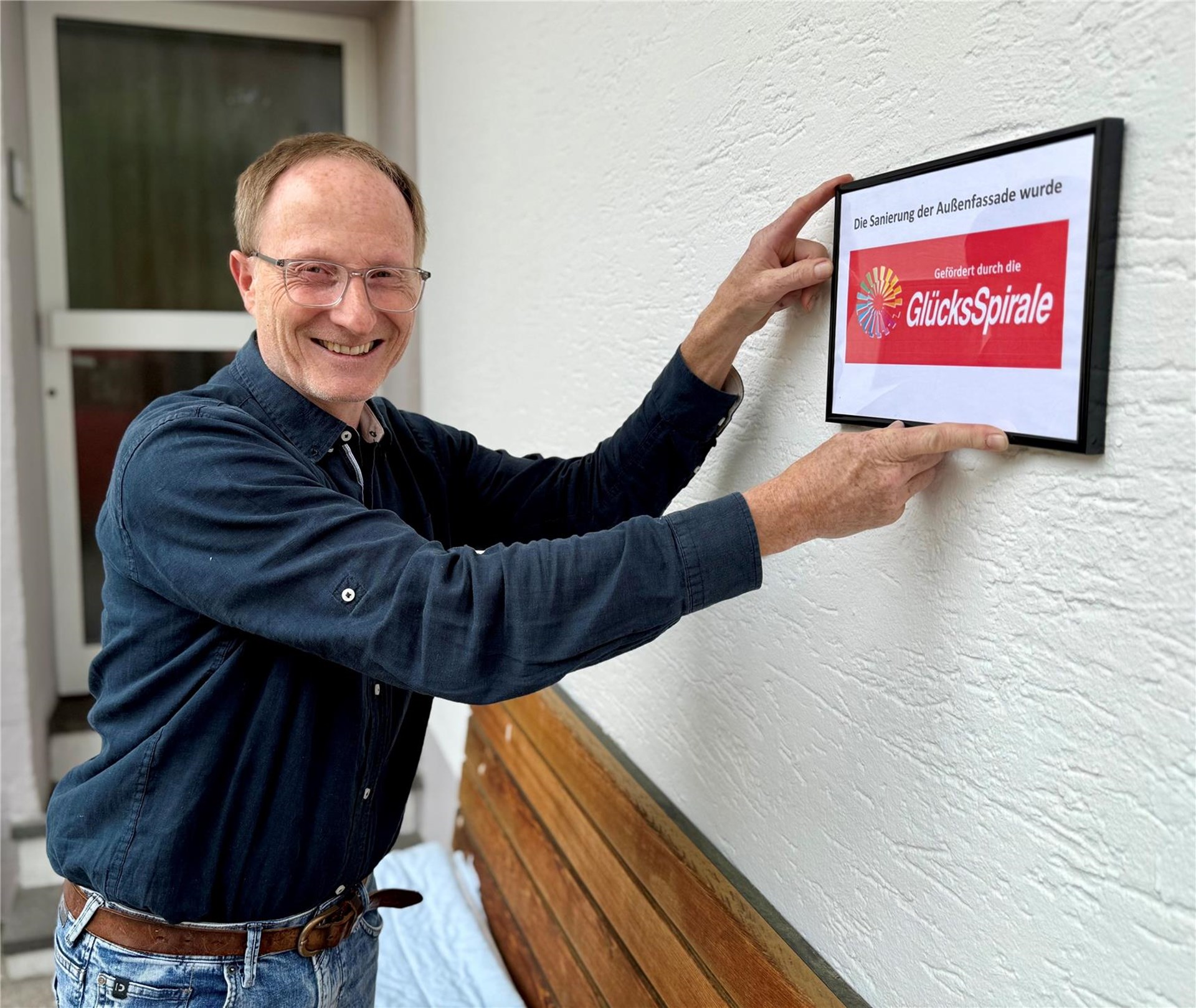 Joachim Grämer, Einrichtungsleiter des Hauses St. Christophorus, bringt am sanierten Bereich des Hauses die Fördertafel der GlücksSpirale an und lacht in die Kamera.