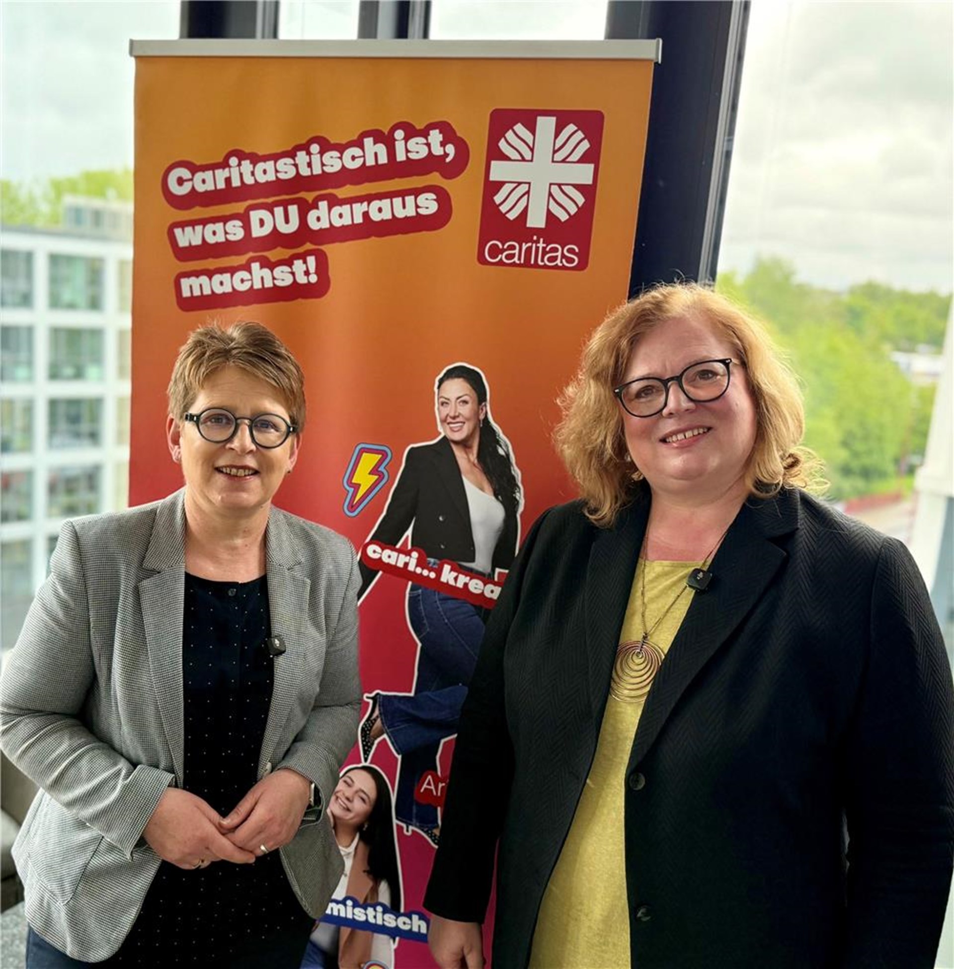 Zwei lächelnde Frauen stehen vor einem orangefarbenen Caritas Roll Up Banner in einem hellen Raum mit großen Fenstern. Auf dem Banner steht „Caritastisch ist, was DU daraus machst!“ neben dem Caritas Logo sowie Bildern weiterer Personen aus der Kampagne.