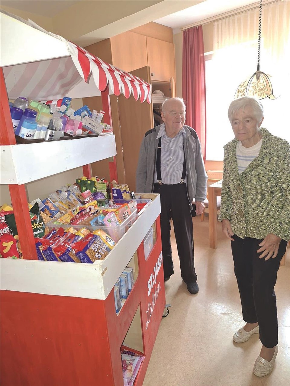 Eine Frau und ein Mann stehen vor einem kleinen Verkaufsstand mit der Aufschrift Sankt Josef Lädchen. Es gibt u.a. Snacks und Drogerieprodukte zu kaufen.