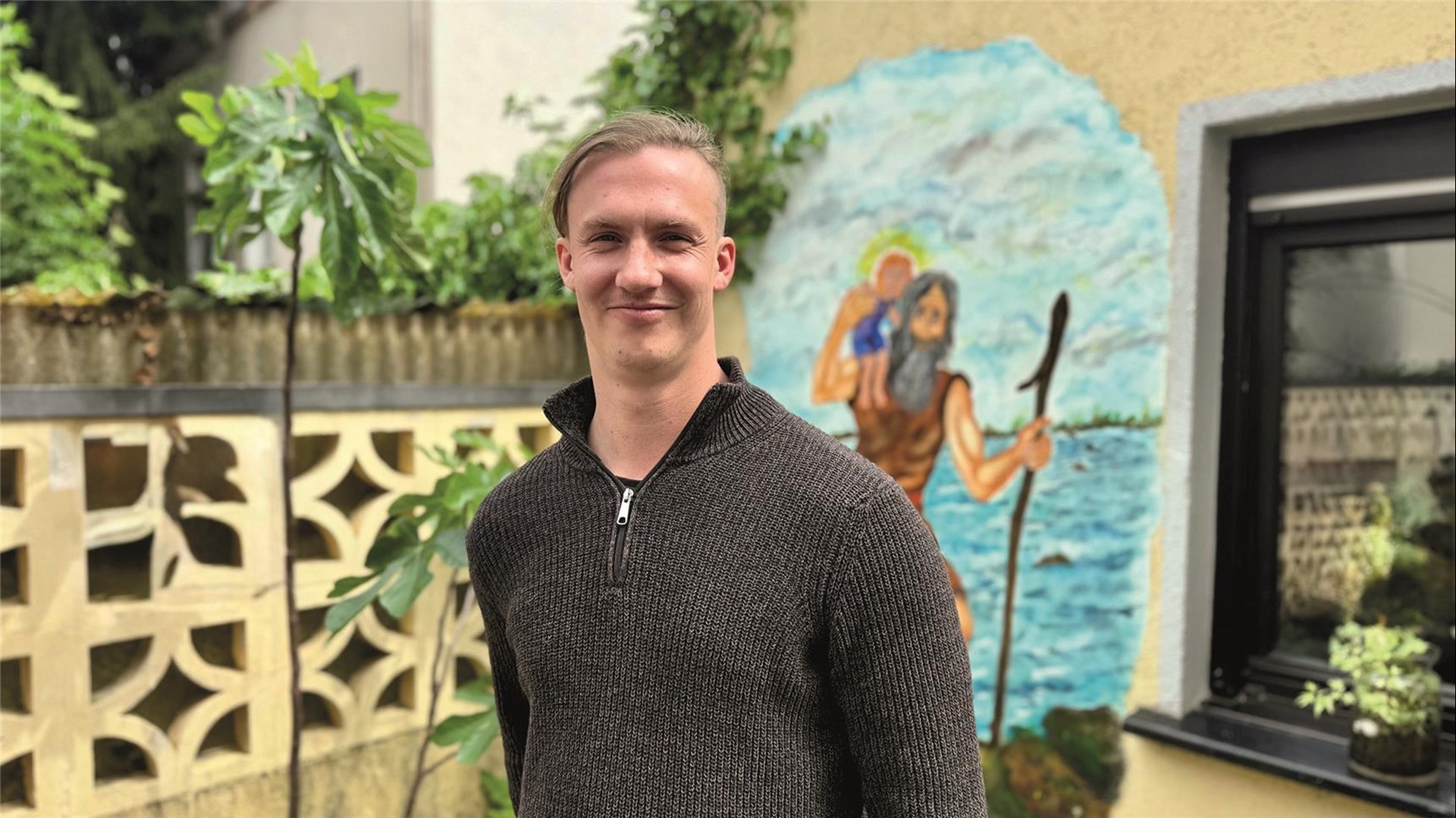Porträt eines lächelnden, jungen Mannes mit kurzen blonden Haaren im dunklen Strickpullover im Freien. Im Hintergrund ist ein Wandgemälde zu sehen, das den heiligen Christopherus mit dem Jesuskind auf den Schultern darstellt.