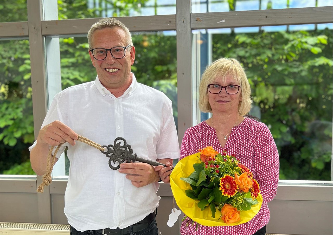 Zwei Personen stehen vor einem großen Fenster. Die Frau hat einen Blumenstrauß in der Hand, der Mann einen großen Schlüssel.