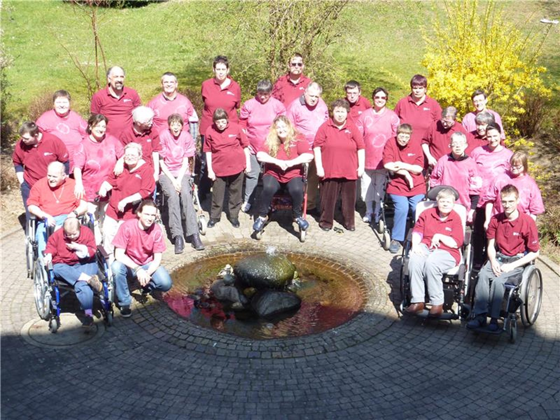 Gruppenfoto von einer Gruppe Frauen und Männer mit rot-pinken Tshirts, die draußen im Halbkreis um einen Brunnen herum stehen oder im Rollstuhl sitzen.