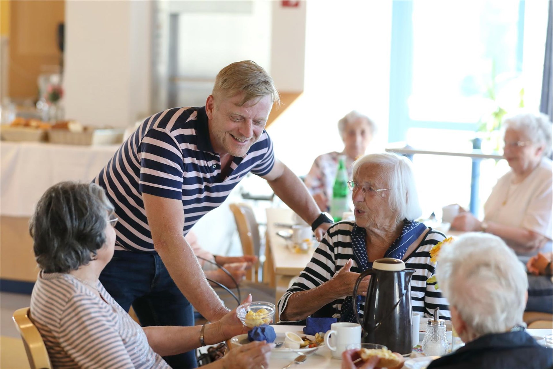 Drei Bewohnerinnen sitzen im Speisesaal beim Frühstück. An ihrem Tisch steht ein Mann und lacht, sie unterhalten sich.