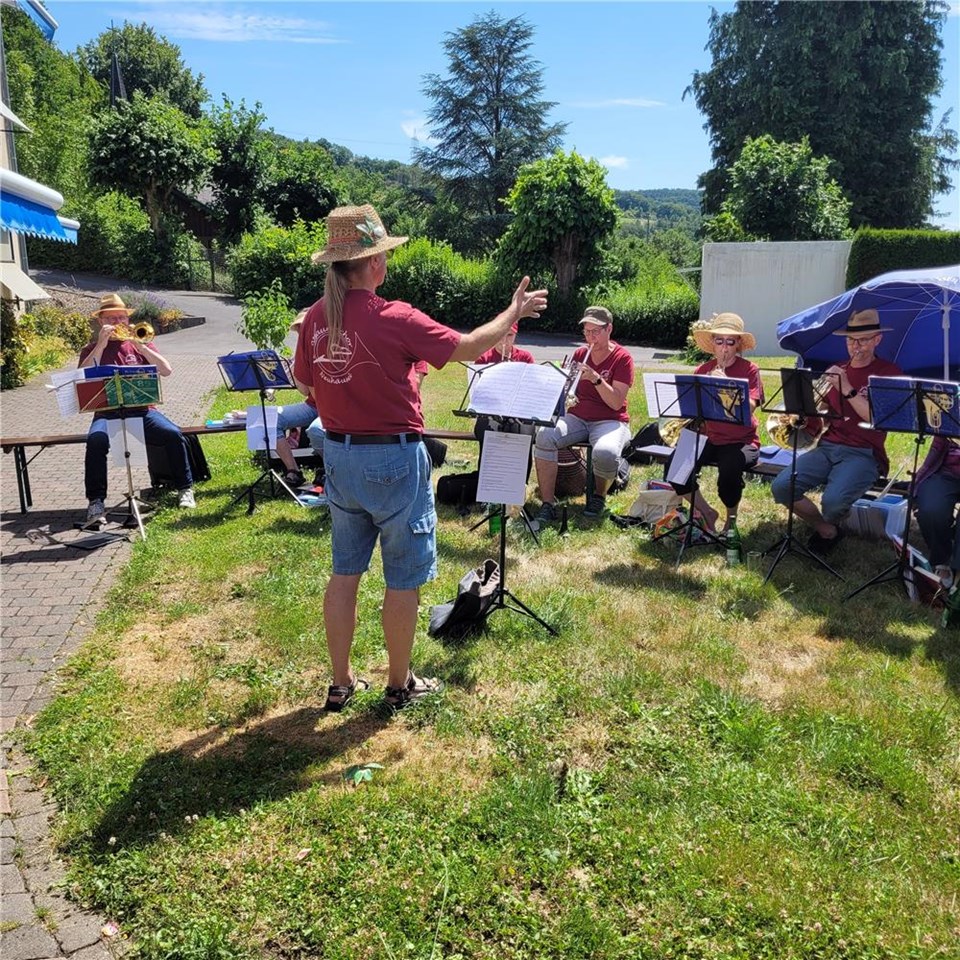 Eine Gruppe von Musikern des evangelischen Posaunenchores Neuhäusel spielt im Garten des Caritas-Altenzentrums St. Josef in Arzbach. Der Dirigent steht mit Strohhut vor den Musikern, die auf Stühlen im Halbkreis sitzen und ihre Blasinstrumente spielen.