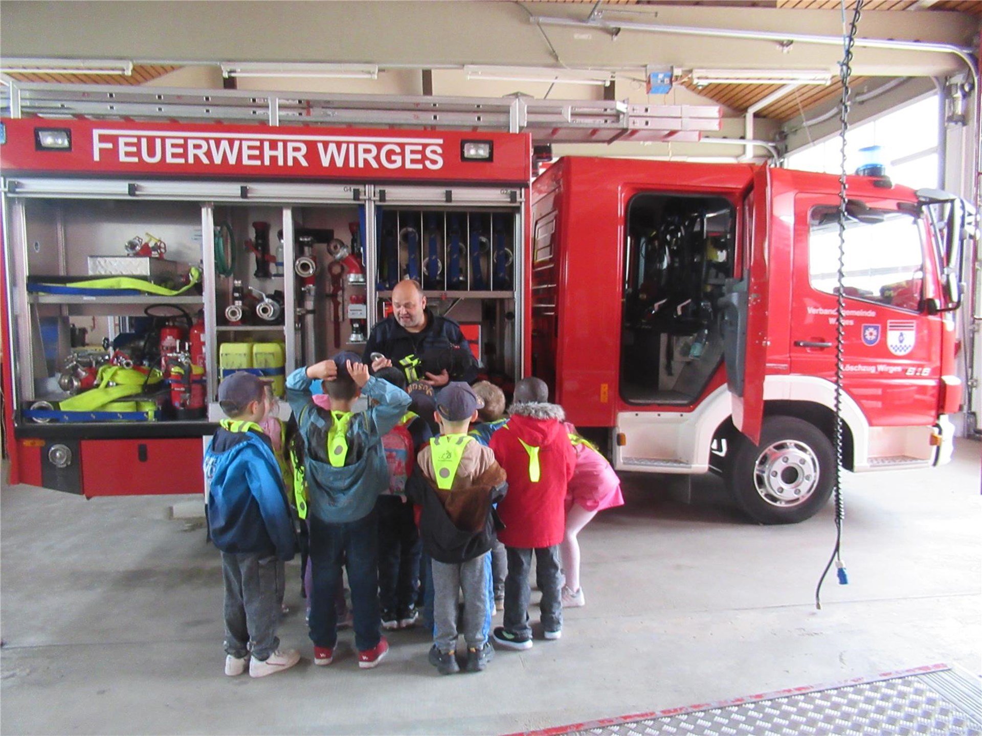 Eine Gruppe von Vorschulkindern der Integrativen Kindertagesstätte St. Franziskus in Wirges steht gemeinsam mit einem Feuerwehrmann vor einem Löschfahrzeug der Freiwilligen Feuerwehr Wirges. Die Kinder erhalten Einblicke in die Ausrüstung und die Aufgaben der Feuerwehr, während das geöffnete Einsatzfahrzeug im Hintergrund zu sehen ist.