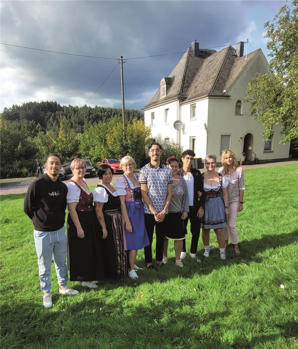 Eine Gruppe von Männern und Frauen steht auf einer Wiese vor einem Haus. Die Frauen tragen Dirndl.