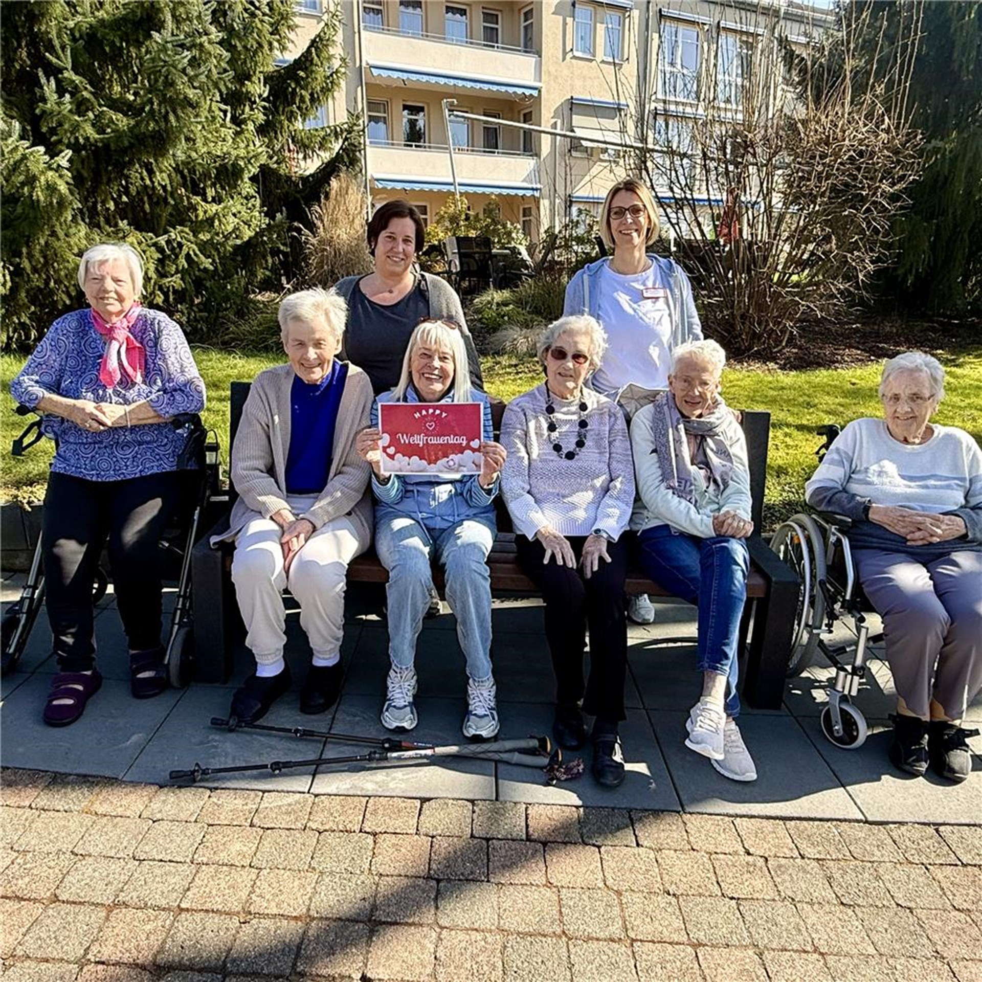 Eine Gruppe Frauen sitzt im Garten und präsentiert ein Schild mit der Aufschrift "Happy Weltfrauentag".